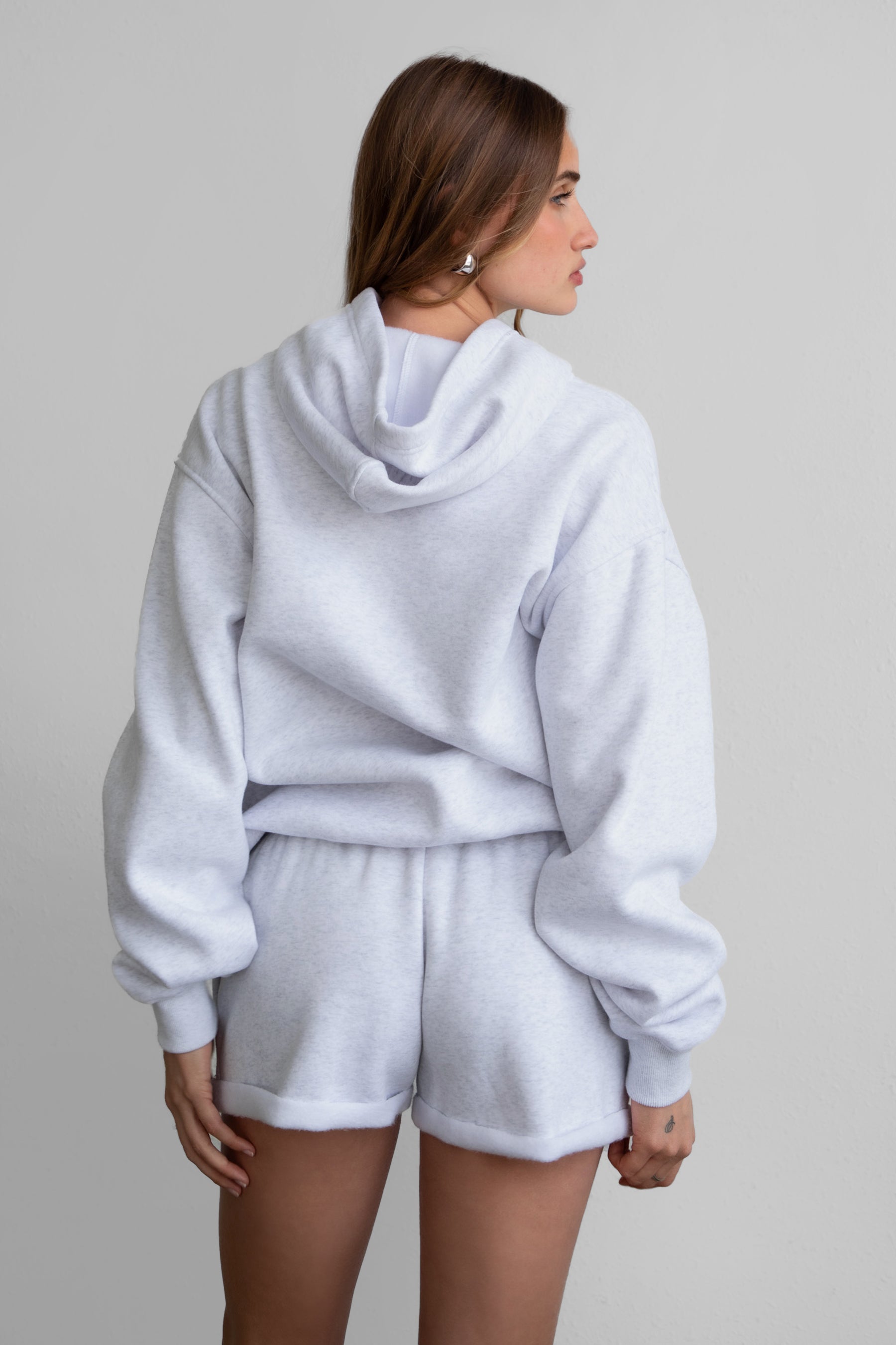 A female model wearing a heather ash white oversized hoodie with a hood, kangaroo pocket, and ribbed cuffs. She is also wearing matching white shorts.