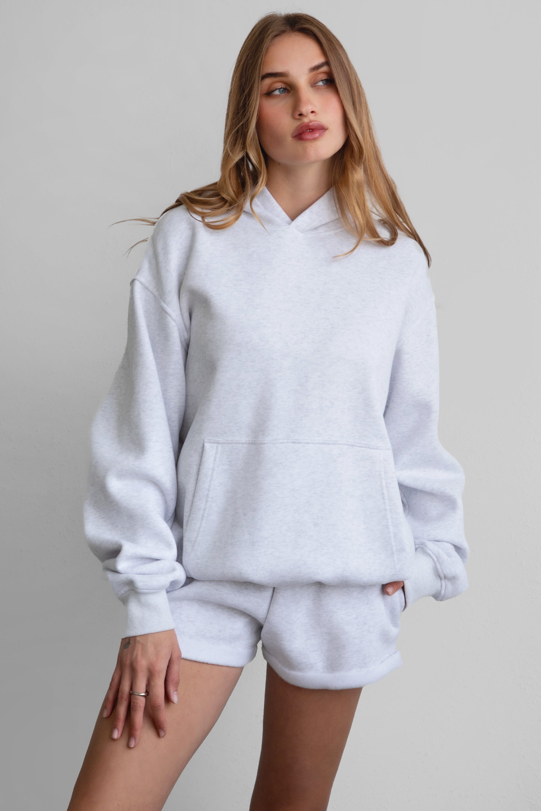 A female model wearing a heather ash white oversized hoodie with a hood, kangaroo pocket, and ribbed cuffs. She is also wearing matching white shorts.