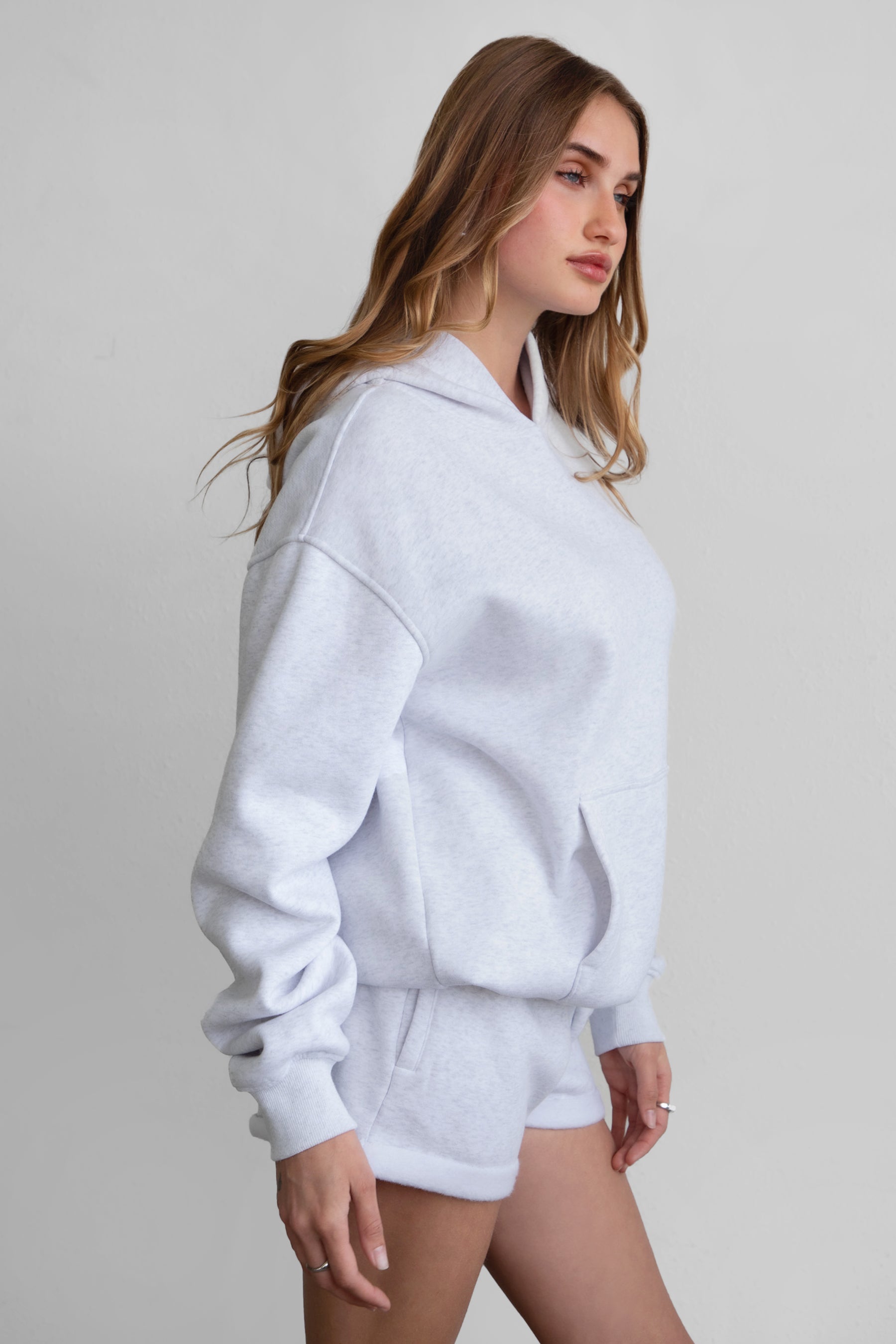 A female model wearing a heather ash white oversized hoodie with a hood, kangaroo pocket, and ribbed cuffs. She is also wearing matching white shorts.