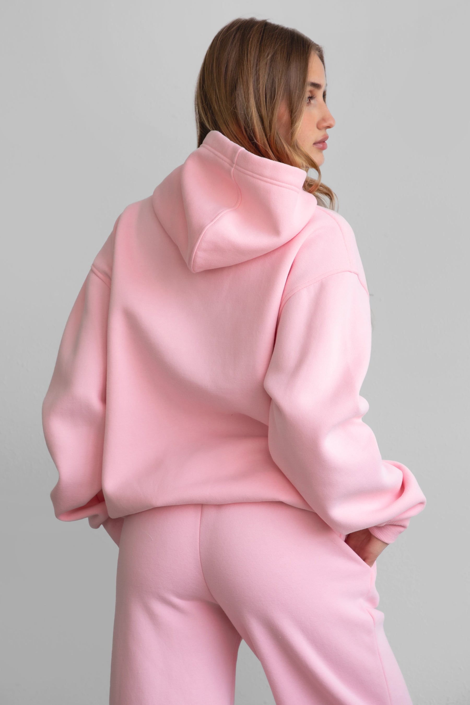A woman wearing a pink oversized hoodie with a kangaroo pocket, matching pants, standing against a plain background.