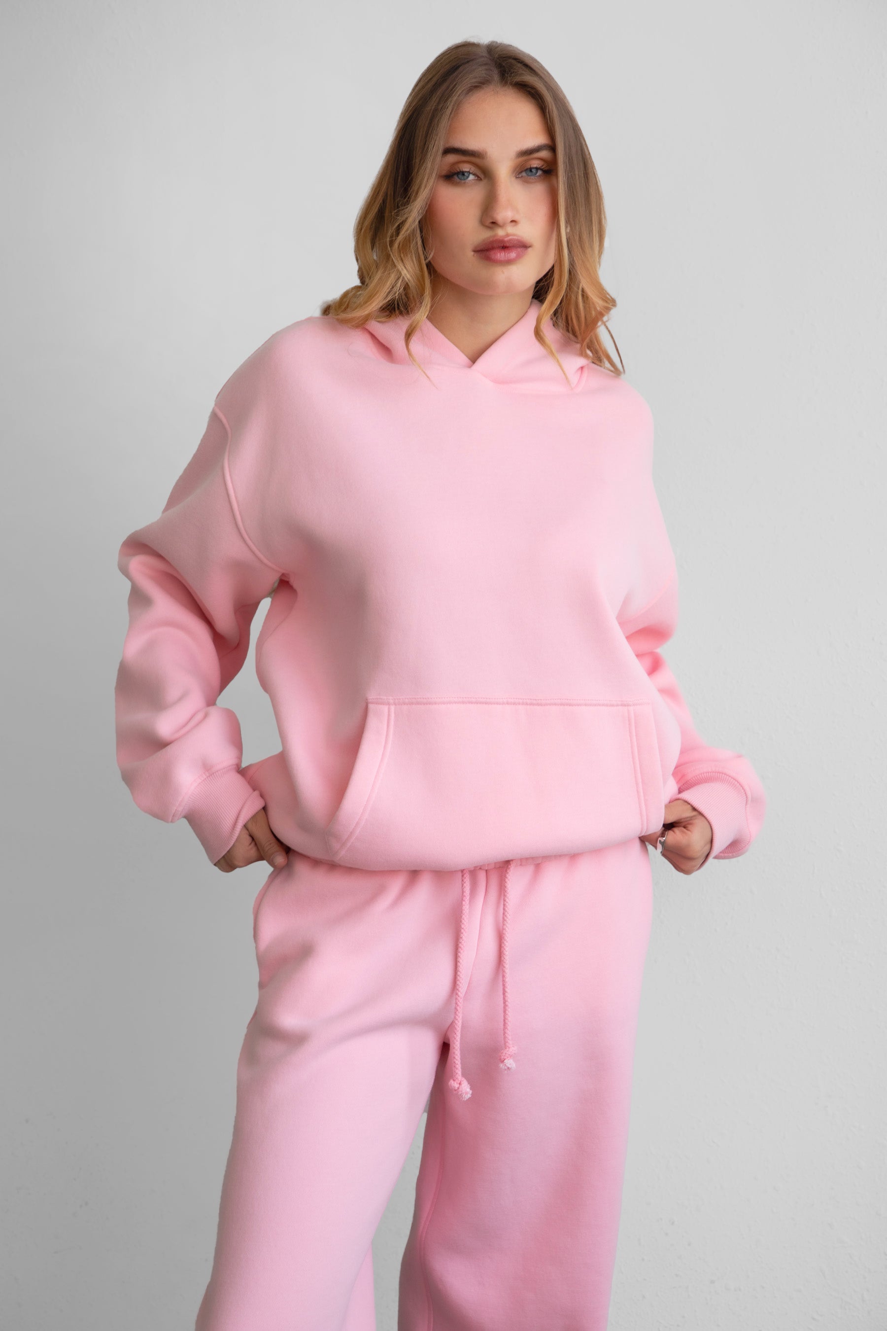 A woman wearing a pink oversized hoodie with a kangaroo pocket, matching pants, standing against a plain background.