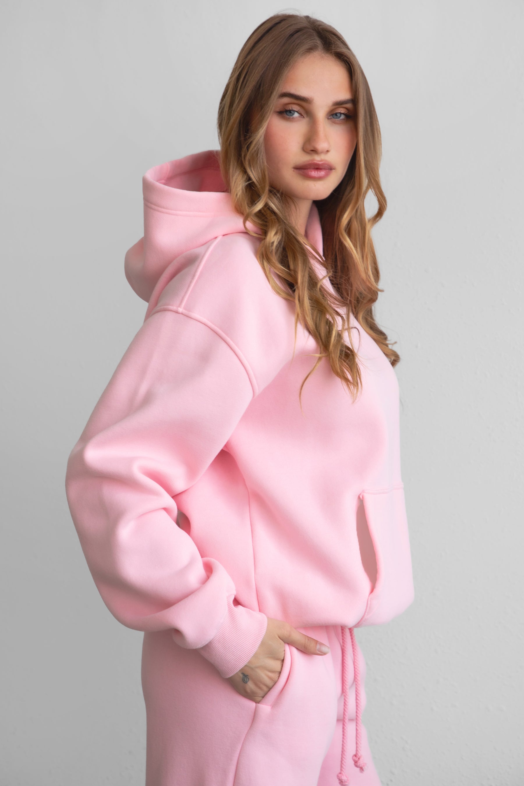 A woman wearing a pink oversized hoodie with a kangaroo pocket, matching pants, standing against a plain background.