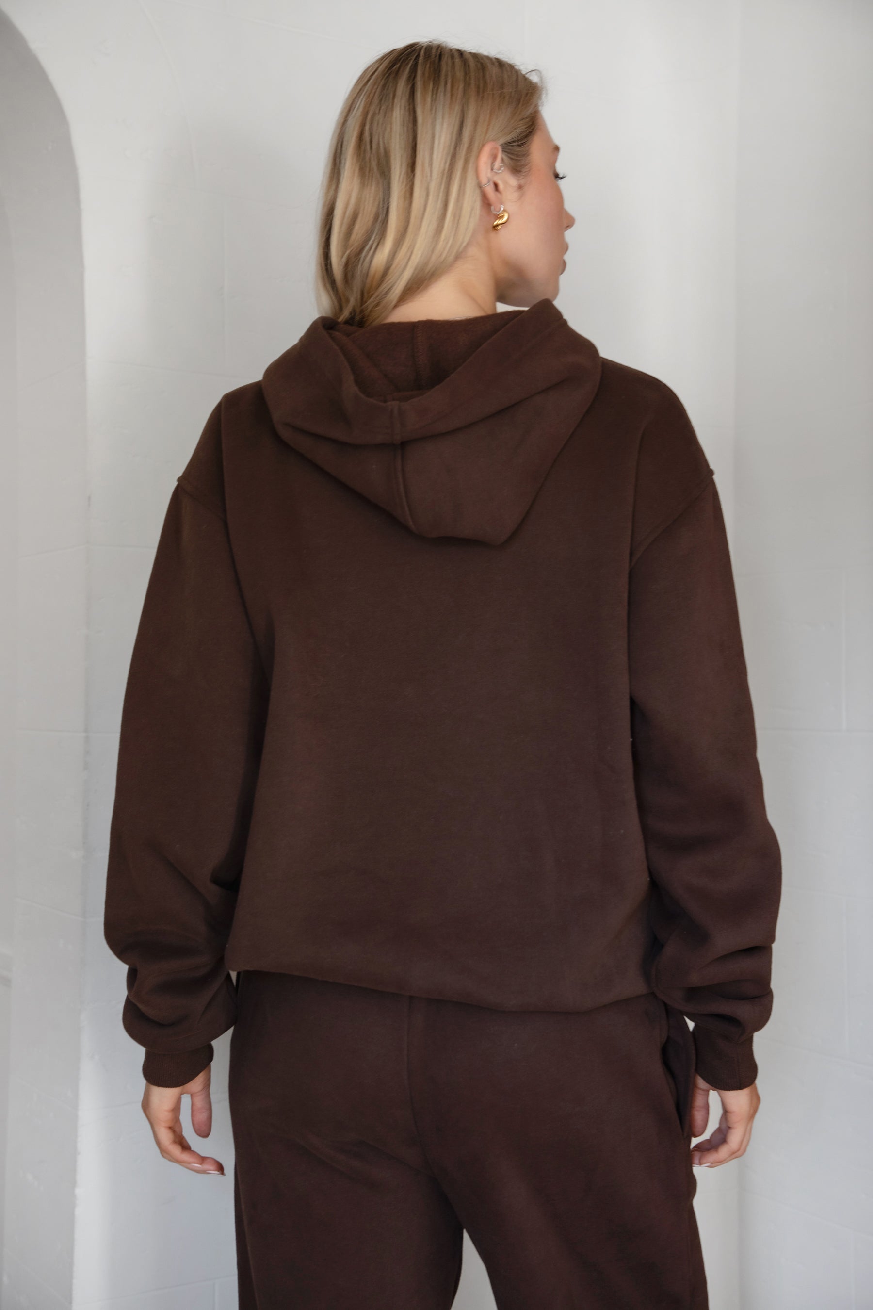 Person wearing a brown hoodie and pants against a white wall