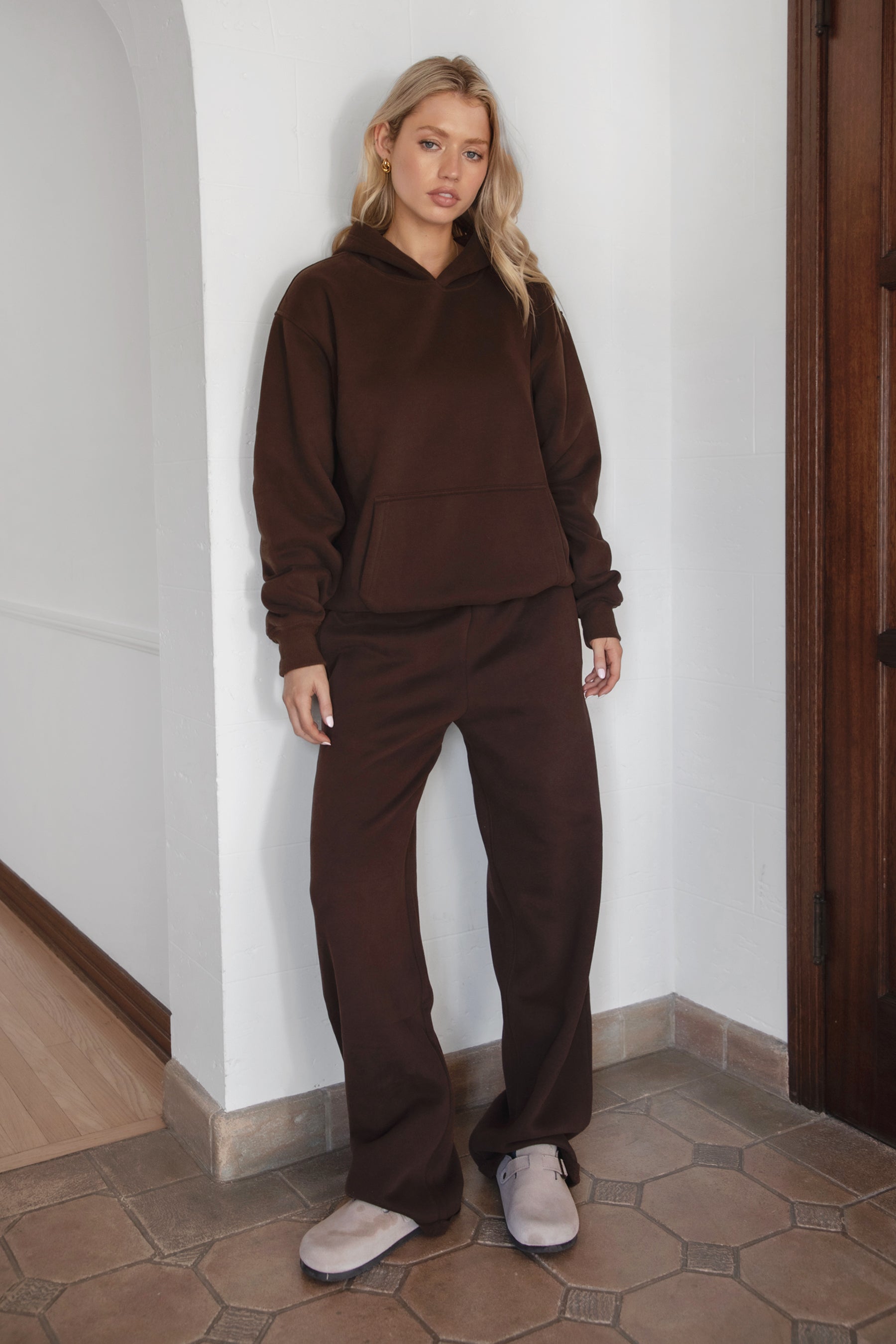 Woman wearing a brown tracksuit standing against a white wall.