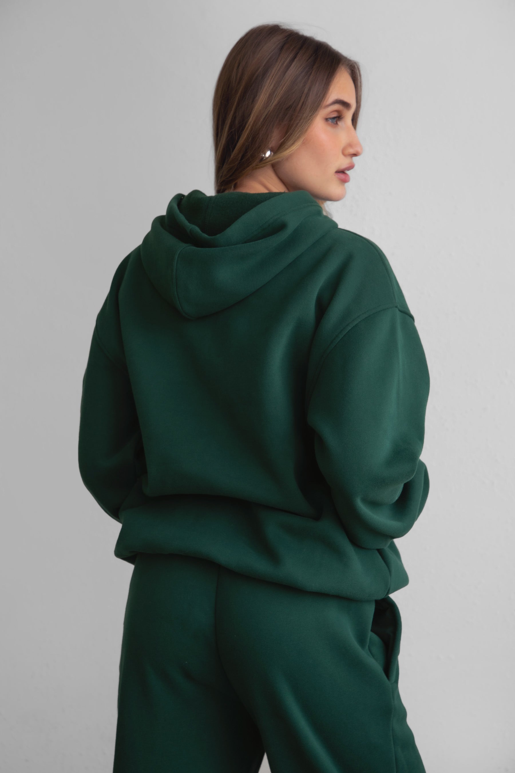A female model wearing a forest green oversized hoodie with a hood, kangaroo pocket, and ribbed cuffs. She is also wearing matching pants. 