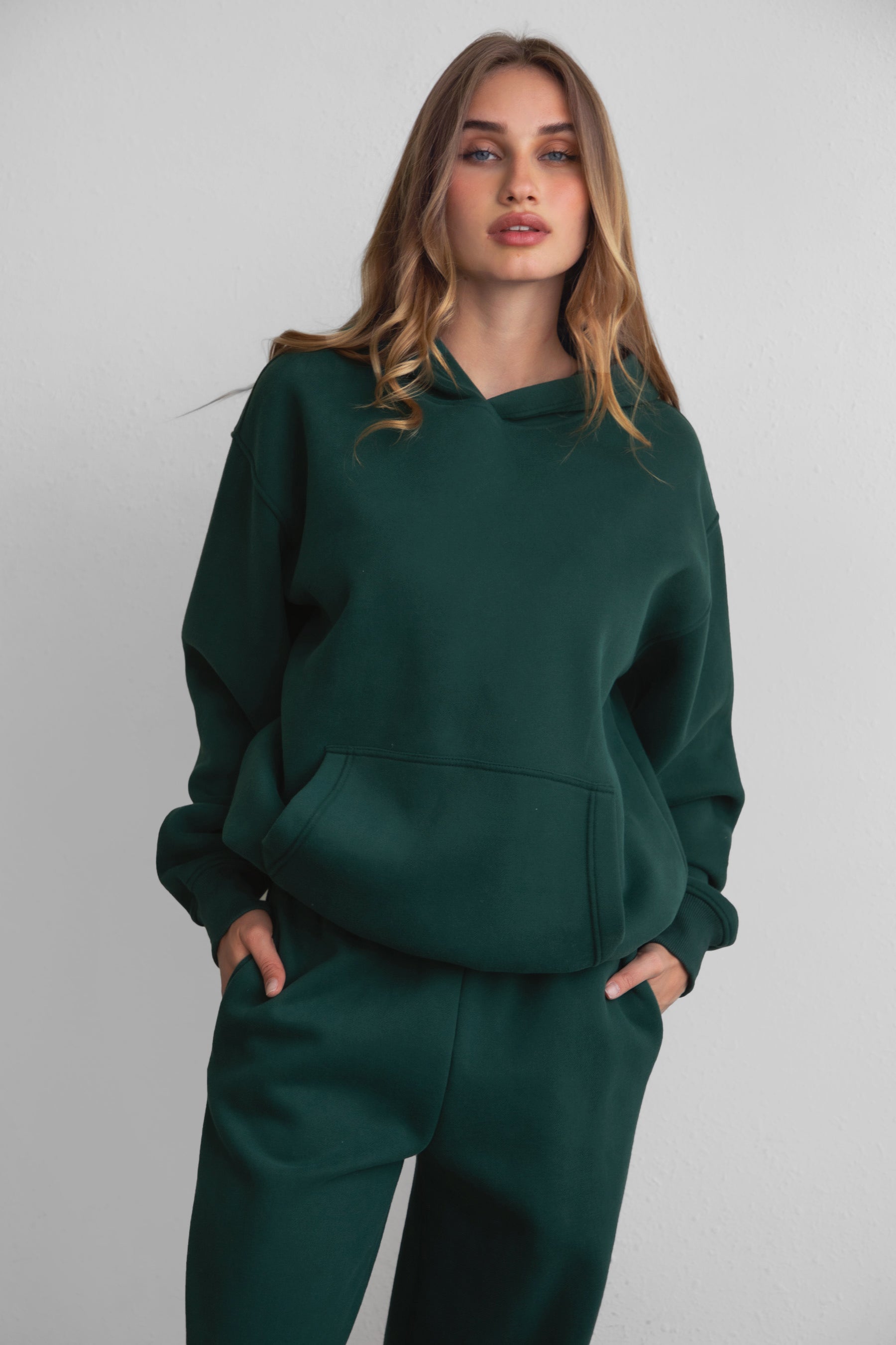 A female model wearing a forest green oversized hoodie with a hood, kangaroo pocket, and ribbed cuffs. She is also wearing matching pants. 