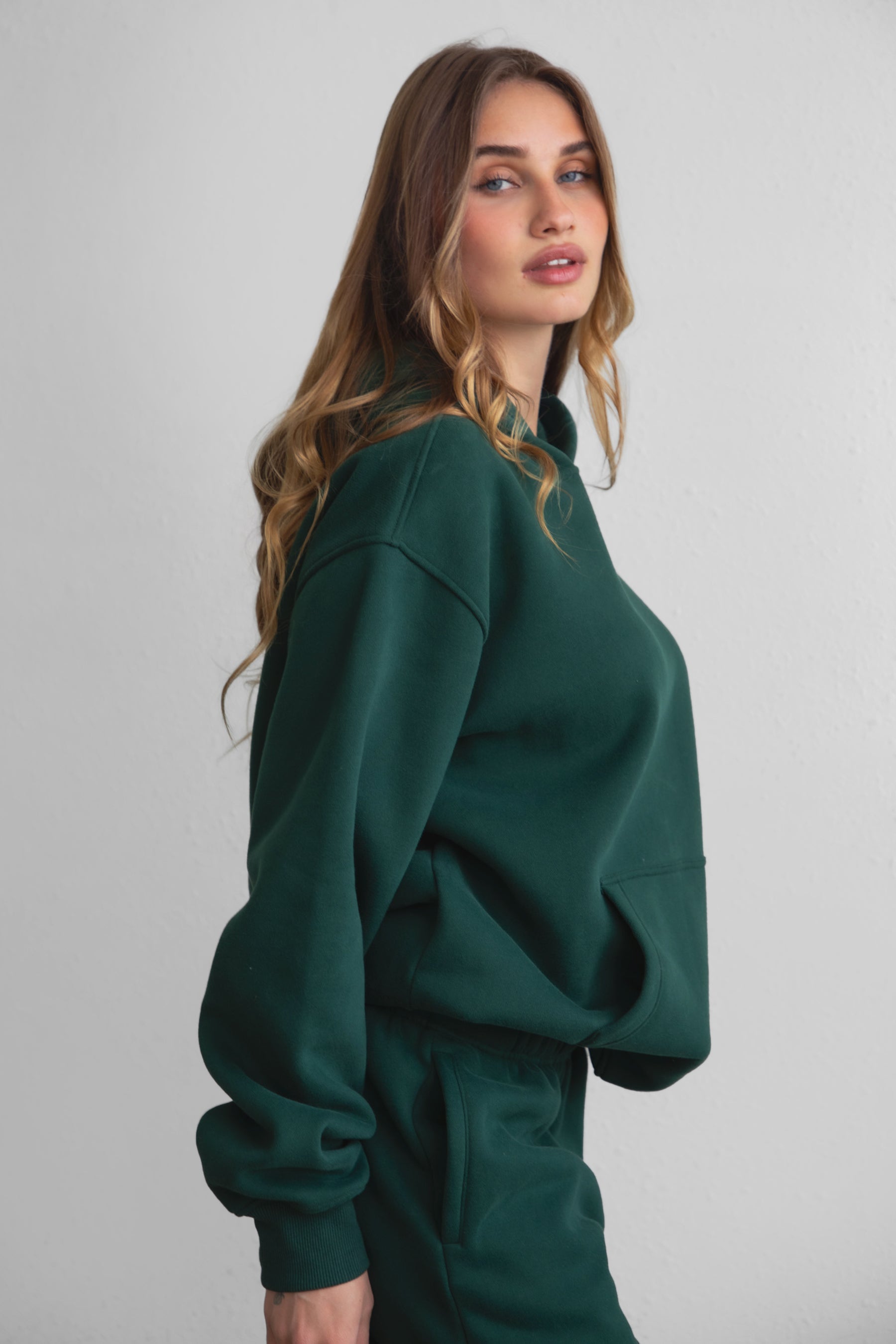 A female model wearing a forest green oversized hoodie with a hood, kangaroo pocket, and ribbed cuffs. She is also wearing matching pants. 