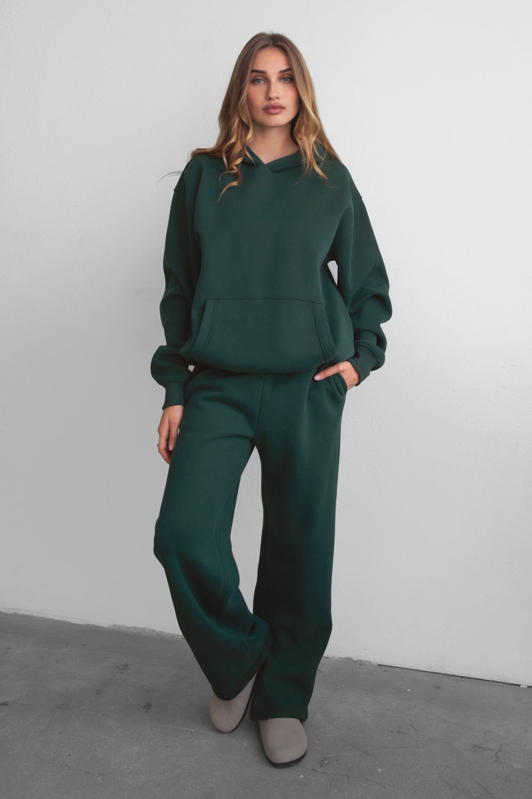 A female model wearing a forest green oversized hoodie with a hood, kangaroo pocket, and ribbed cuffs. She is also wearing matching pants. 