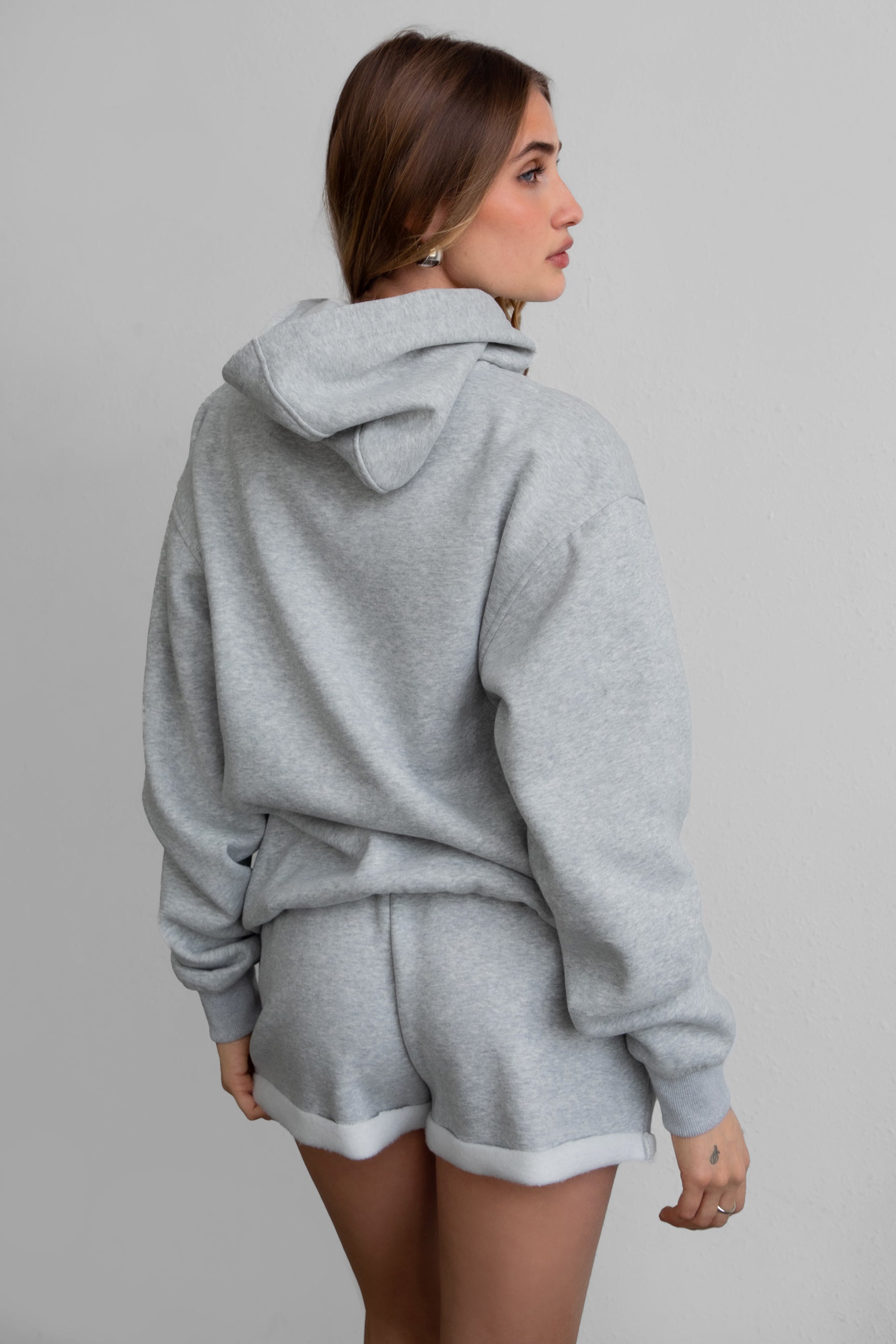 A female model wearing a grey oversized hoodie with a hood, kangaroo pocket, and ribbed cuffs. She is also wearing matching grey shorts.