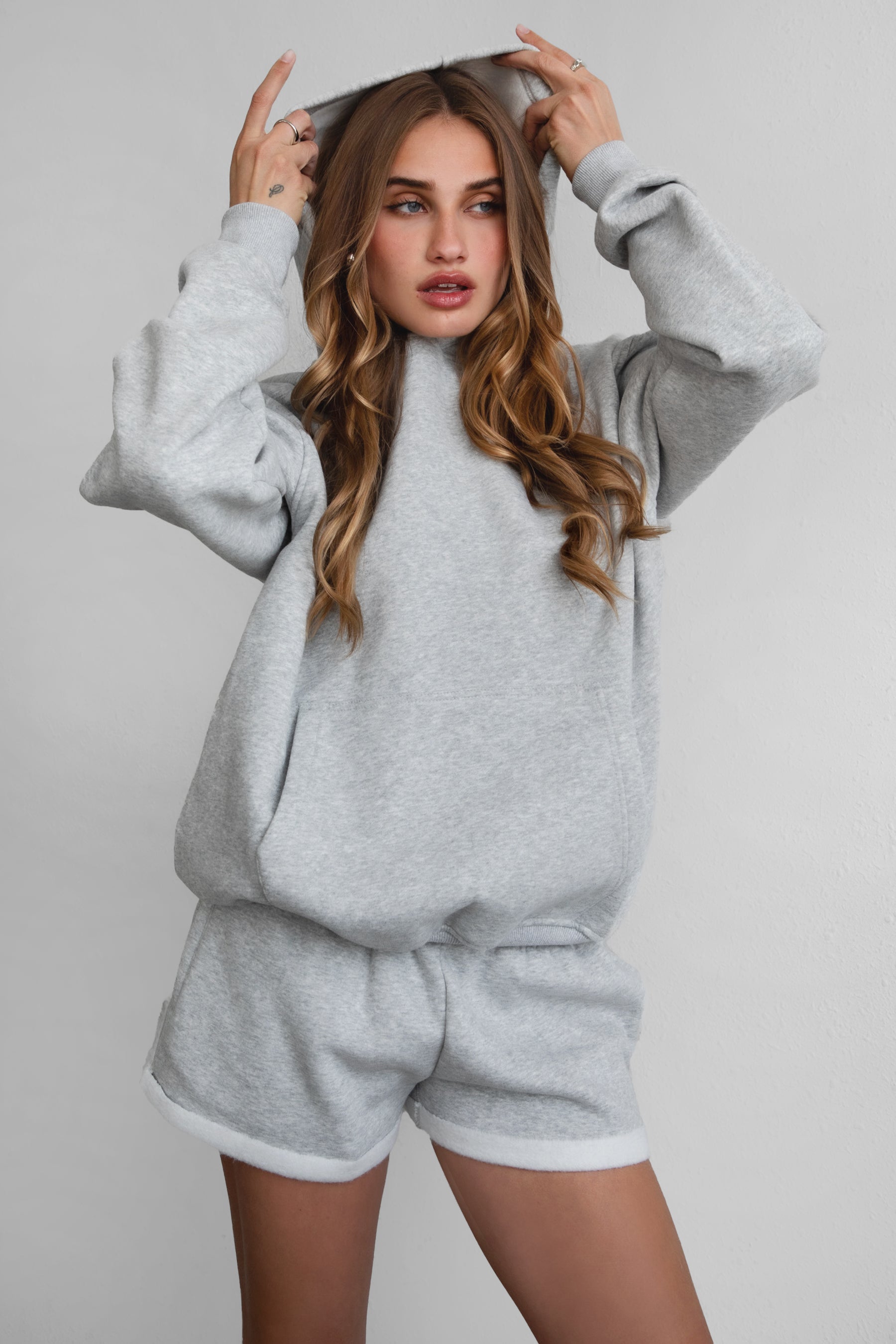 A female model wearing a grey oversized hoodie with a hood, kangaroo pocket, and ribbed cuffs. She is also wearing matching grey shorts.