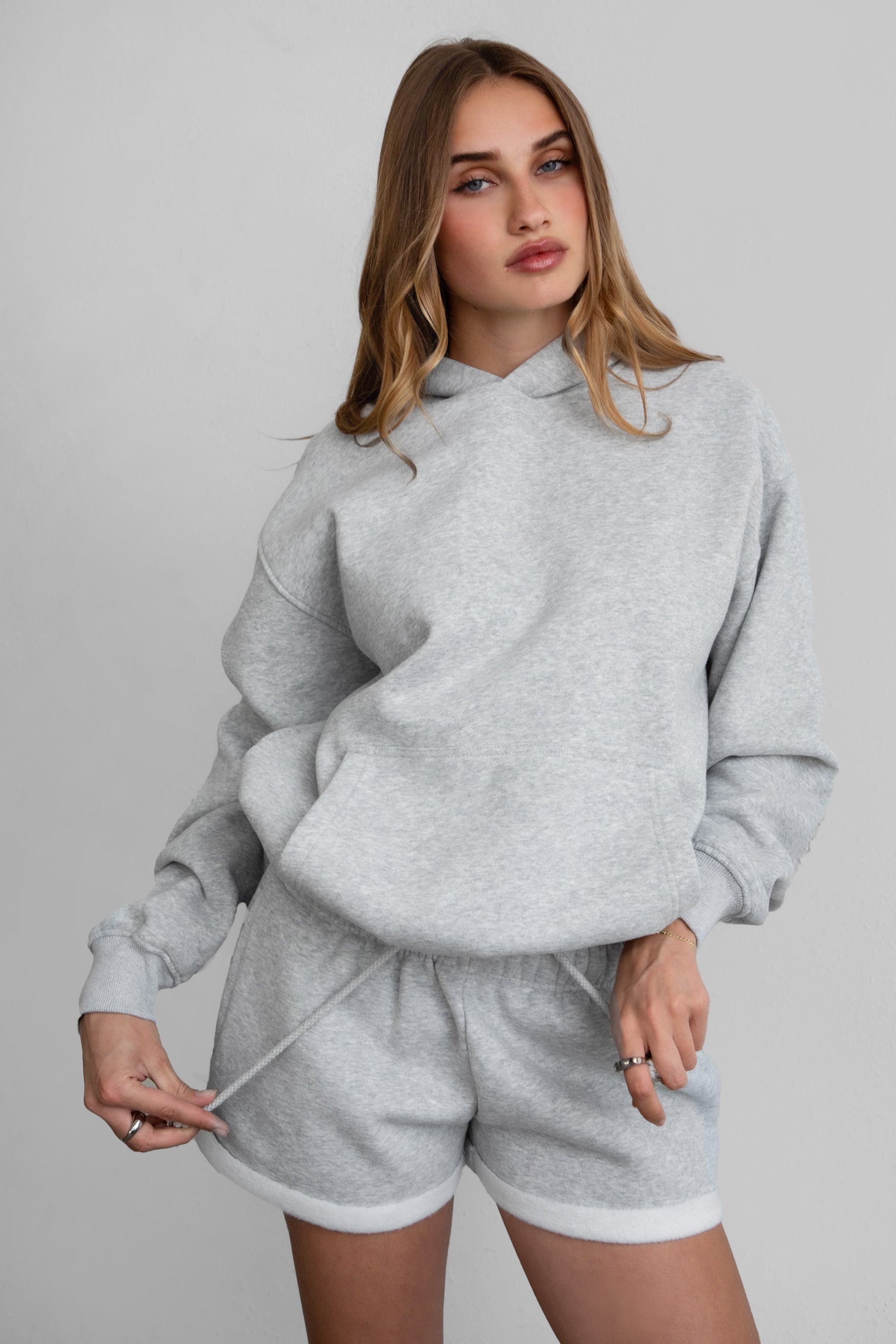 A female model wearing a grey oversized hoodie with a hood, kangaroo pocket, and ribbed cuffs. She is also wearing matching grey shorts.