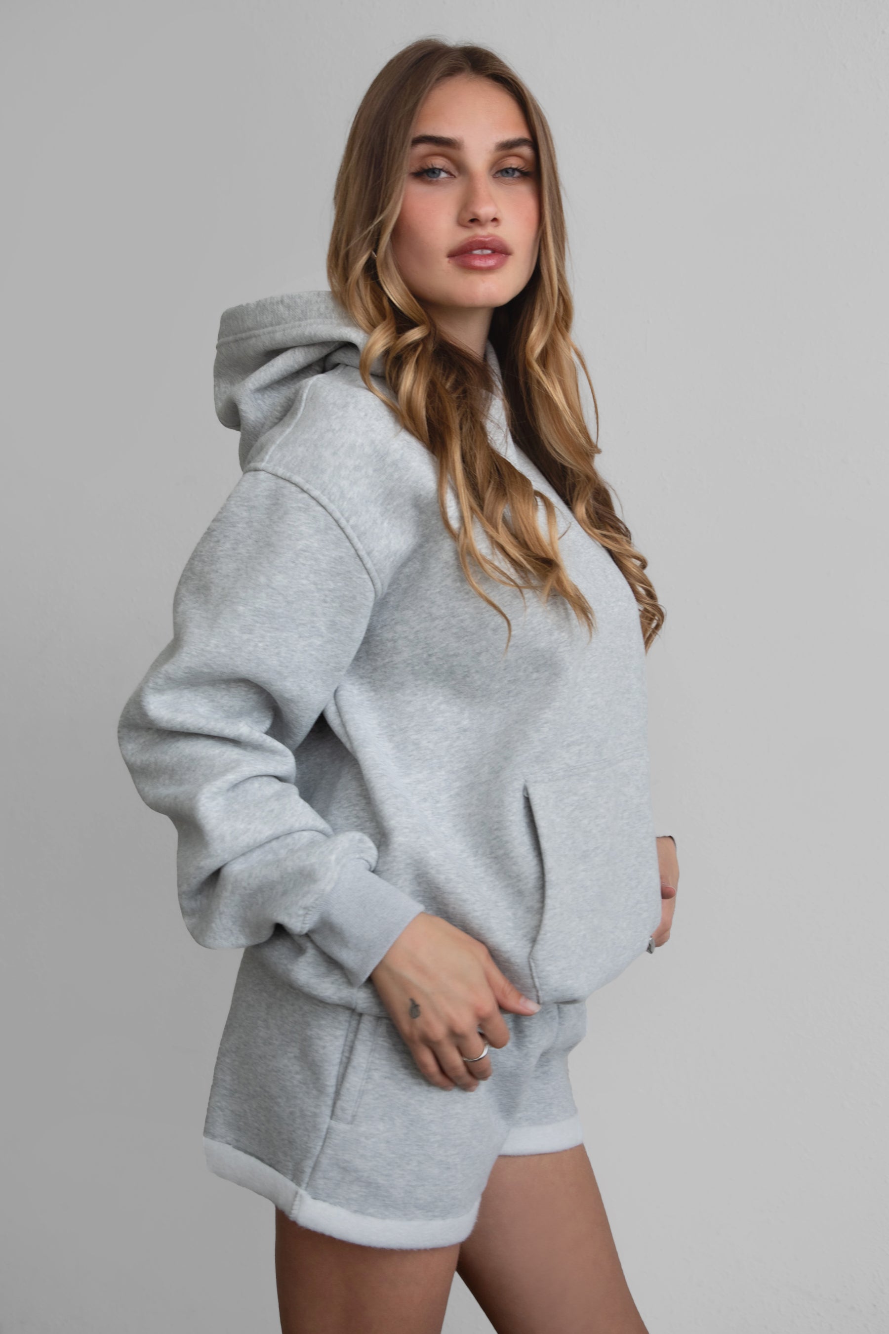 A female model wearing a grey oversized hoodie with a hood, kangaroo pocket, and ribbed cuffs. She is also wearing matching grey shorts.