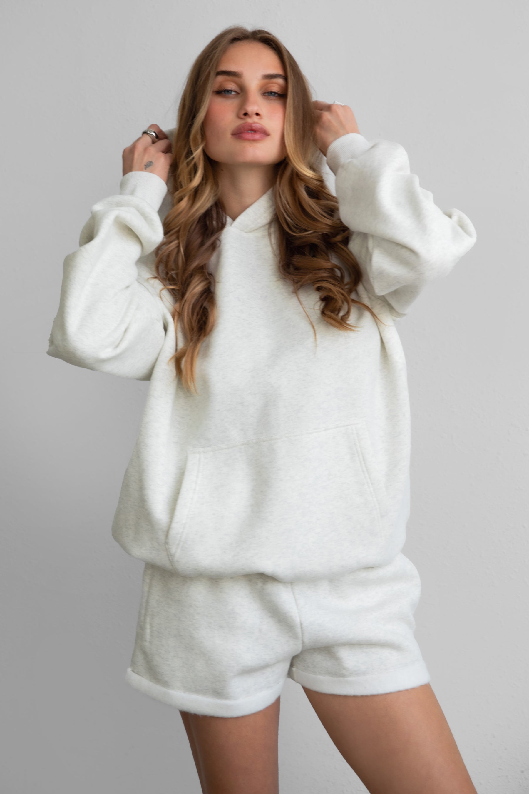 A female model wearing a heather white oversized hoodie with a hood, long sleeves, and standing pose.