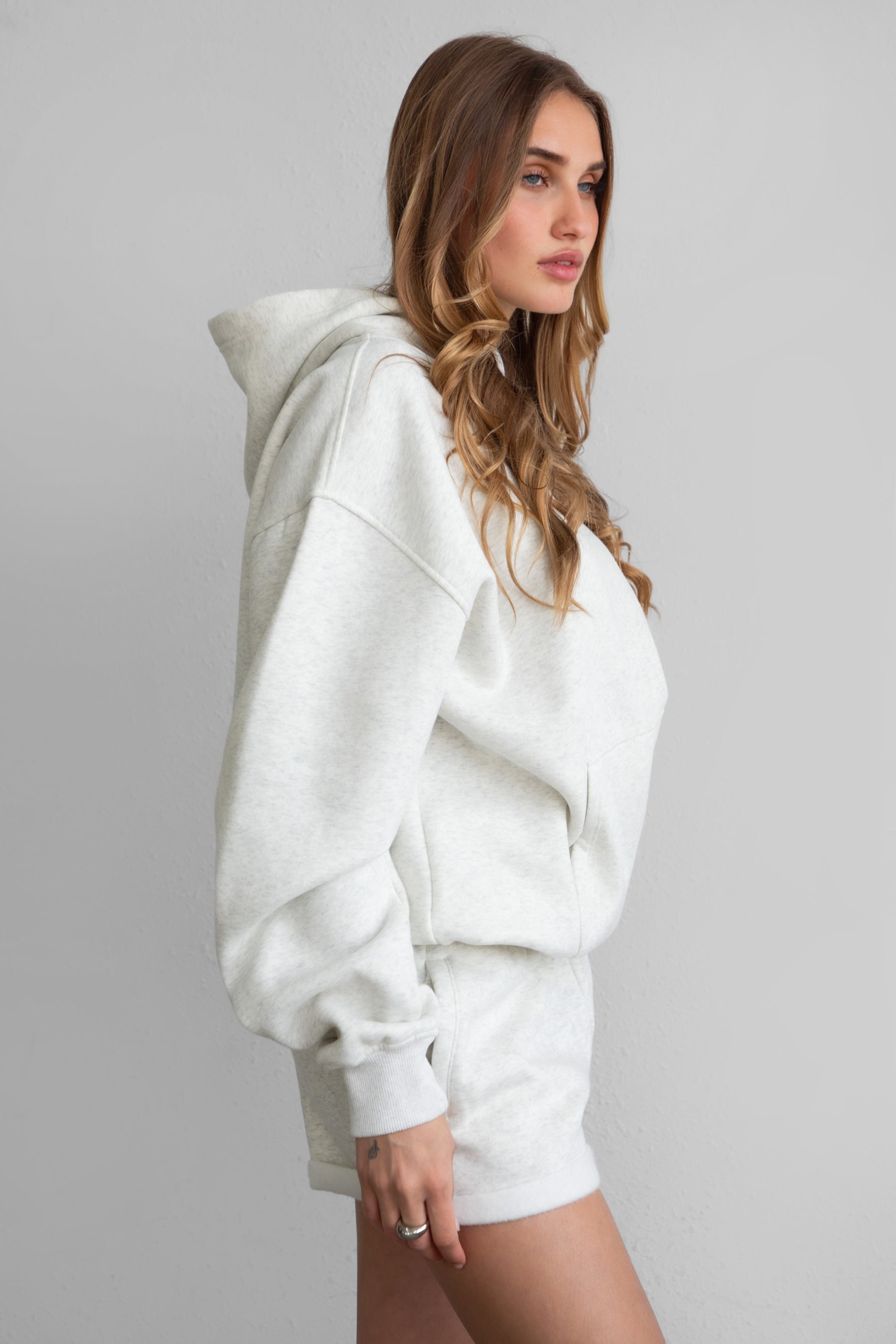 A female model wearing a heather white oversized hoodie with a hood, long sleeves, and standing pose.