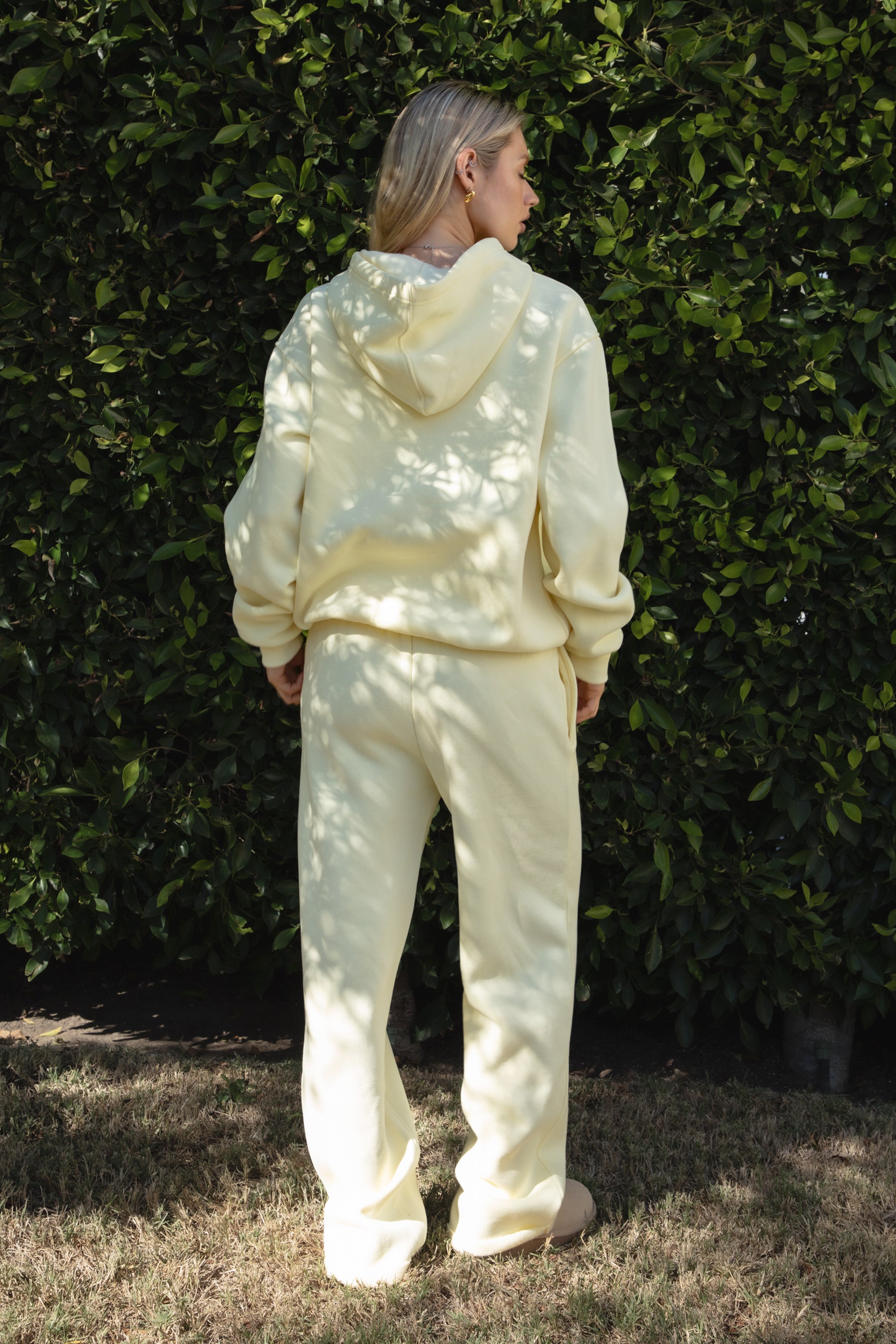 Person wearing a yellow colored hoodie and pants standing against a green hedge.