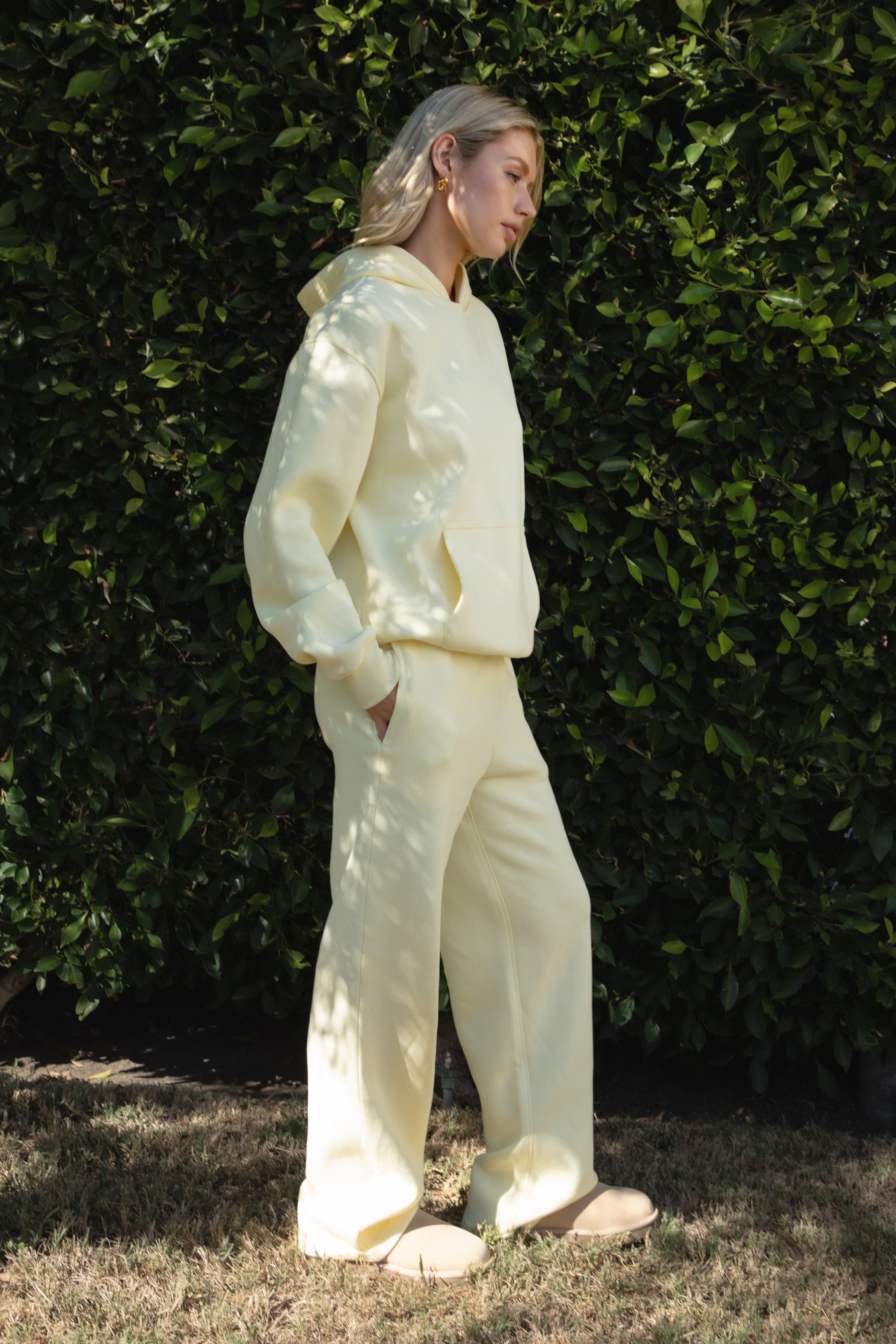 Person wearing a yellow colored hoodie and pants standing against a green hedge.