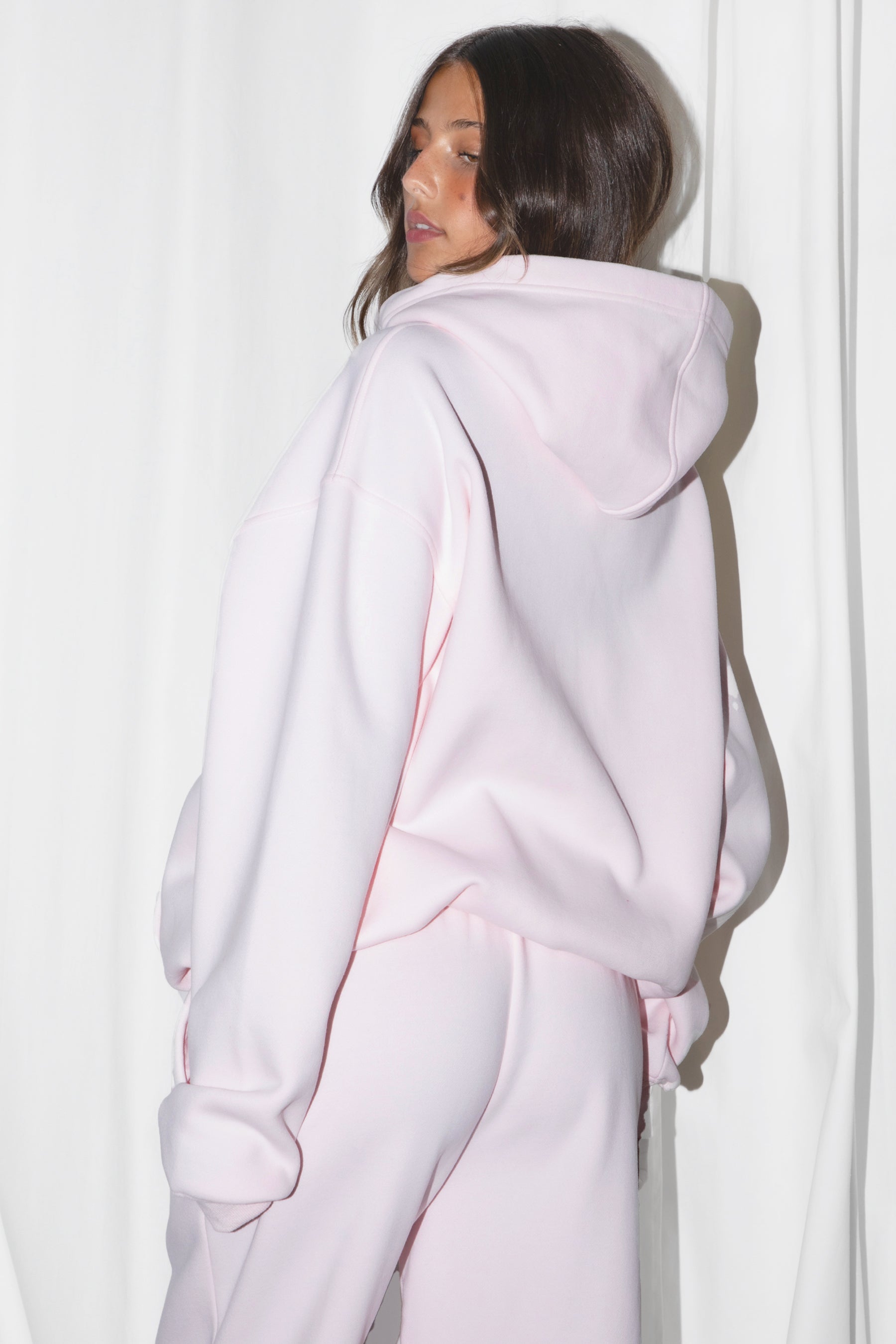 A female model wearing a pale pink oversized hoodie with a hood, long sleeves paired with matching bottoms. 