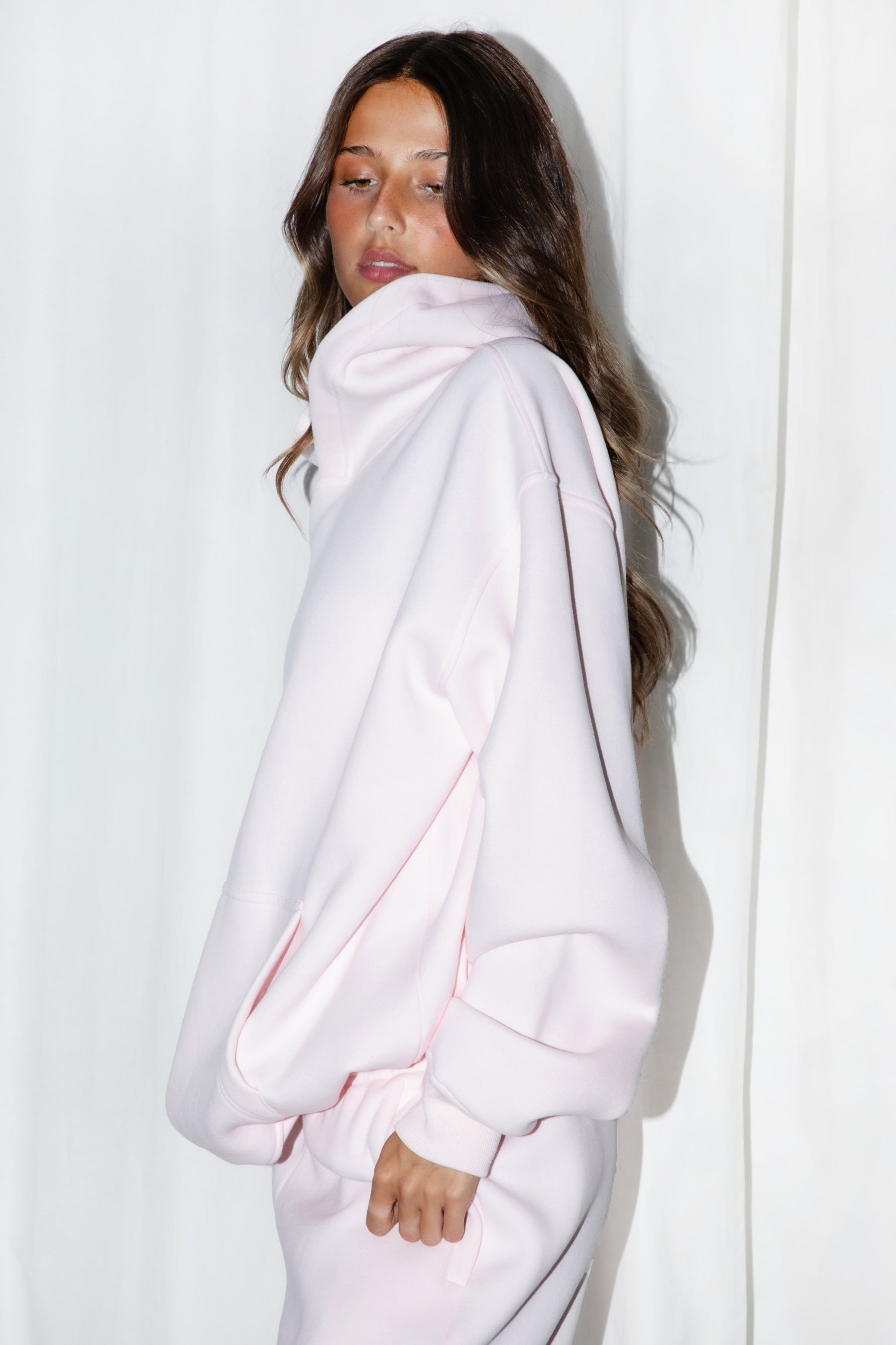 A female model wearing a pale pink oversized hoodie with a hood, long sleeves paired with matching bottoms. 