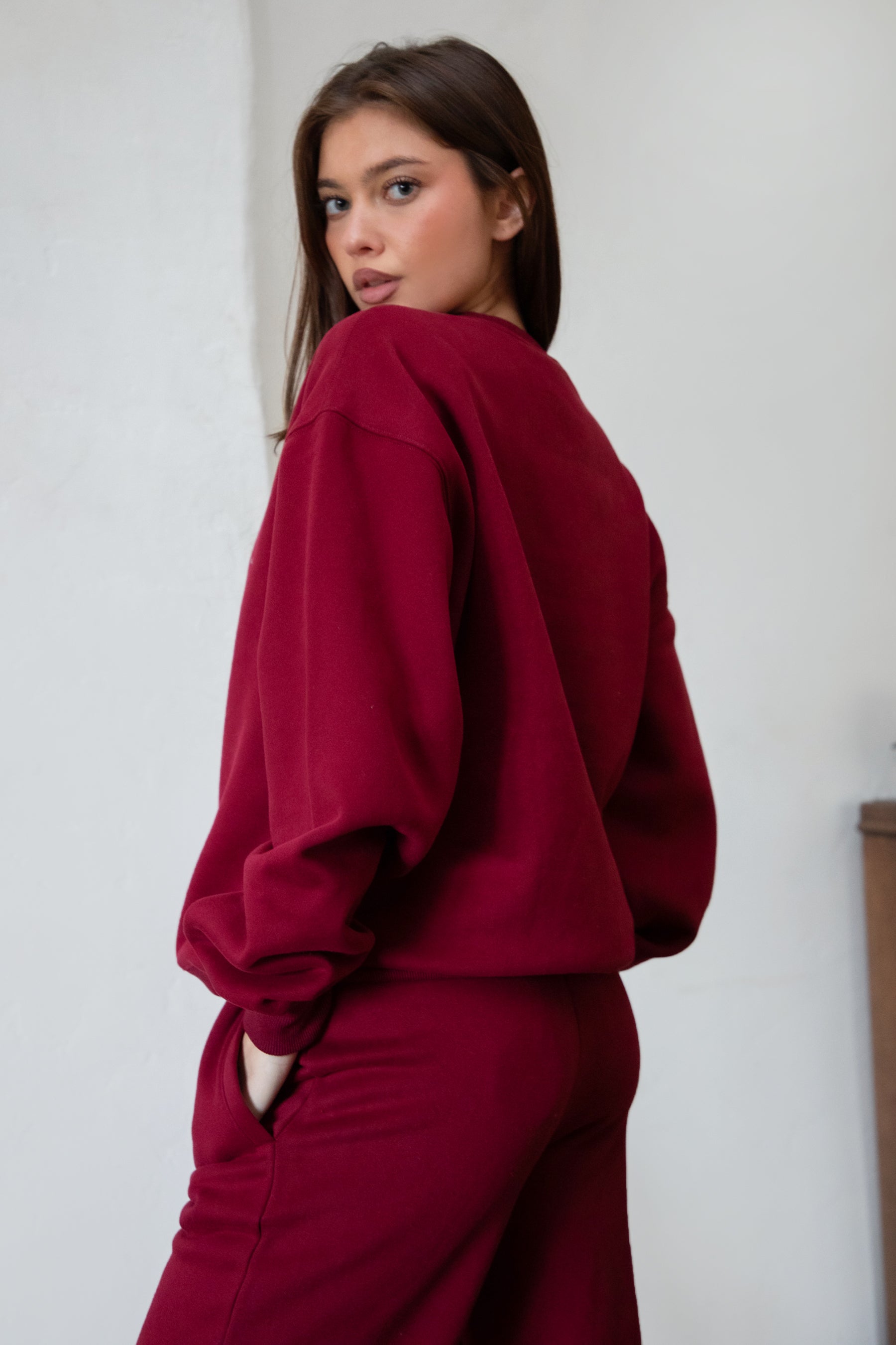 Woman wearing the TIC TOC TJ31656 LANI maroon color fleece crew neck pullover sweatshirt featuring dropped shoulders, ribbed trim, and cozy fleece fabric with the matching bottoms indoors. 