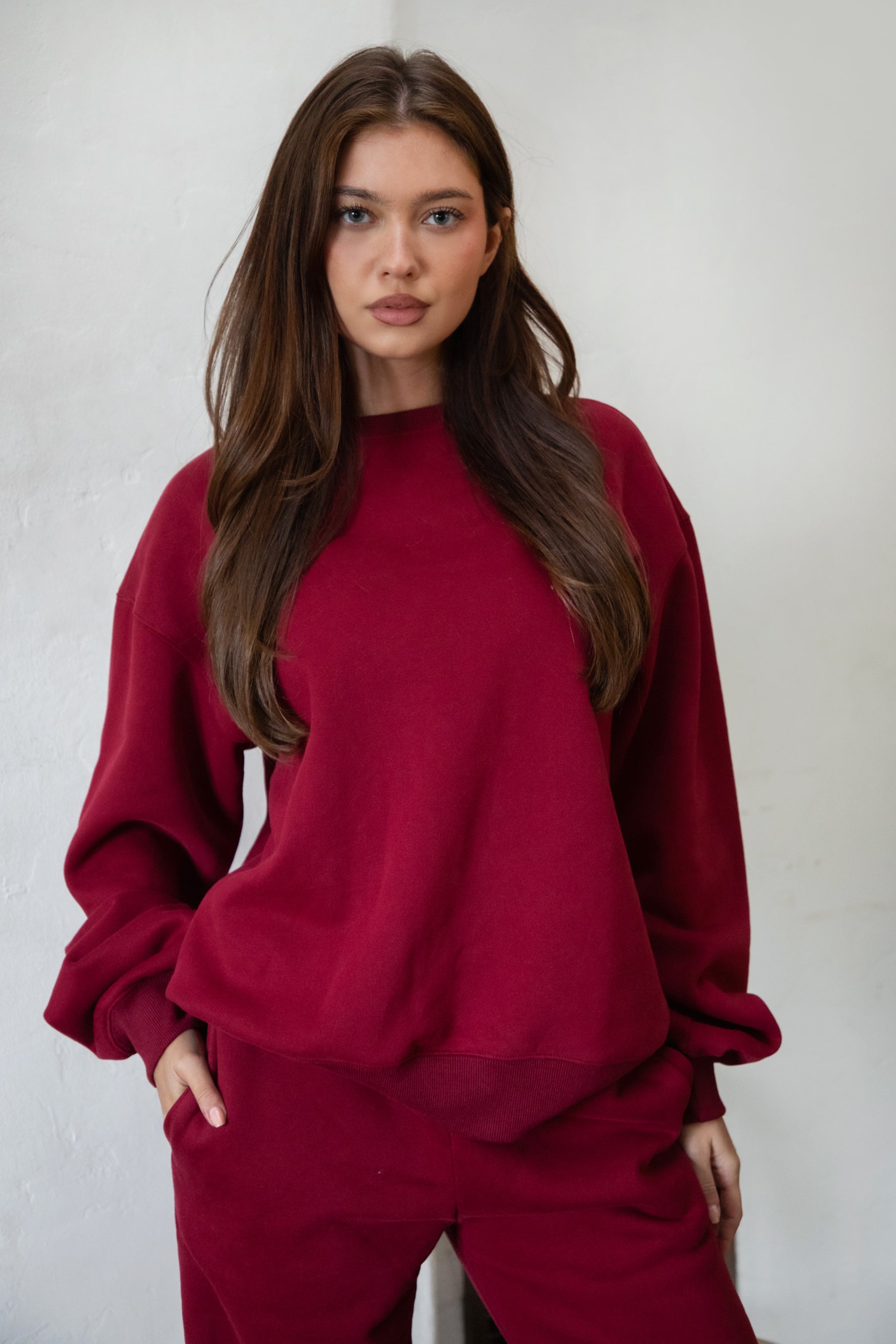 Woman wearing the TIC TOC TJ31656 LANI maroon color fleece crew neck pullover sweatshirt featuring dropped shoulders, ribbed trim, and cozy fleece fabric with the matching bottoms indoors. 