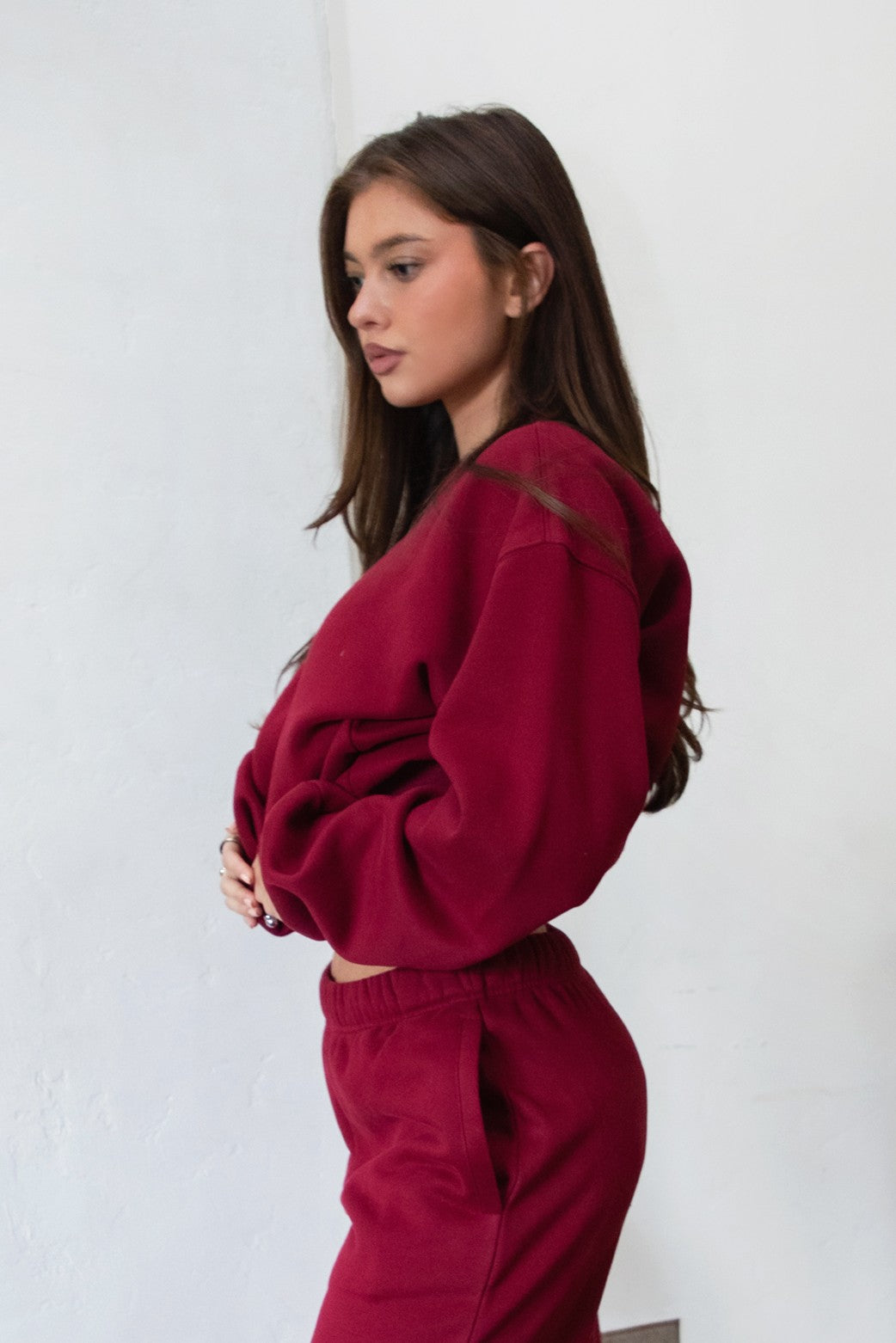 Woman wearing the TIC TOC TJ31656 LANI maroon color fleece crew neck pullover sweatshirt featuring dropped shoulders, ribbed trim, and cozy fleece fabric with the matching bottoms indoors. 